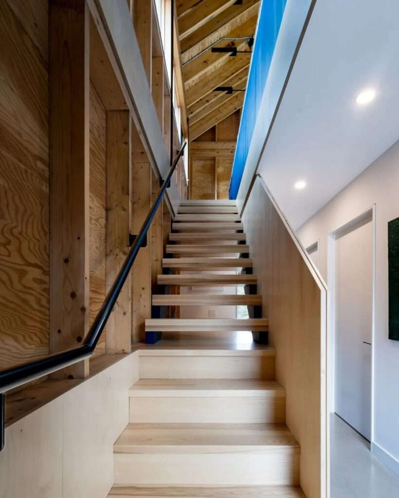 Wooden interior staircase with plywood wall panels and steel handrail connecting the lower private bedrooms to the upper living level.