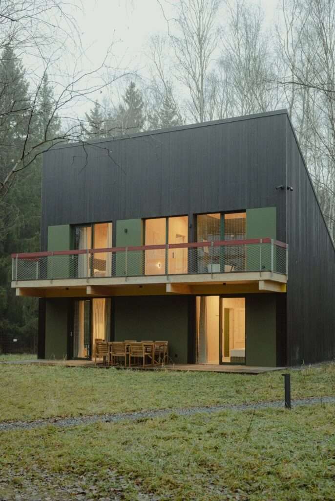 Evening view of a two-story dark timber cabin with warm glowing lights inside large glass windows and doors.