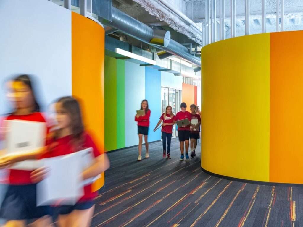 Students carrying laptops along a curved yellow and orange wall pathway, with exposed ductwork overhead and carpeted flooring.