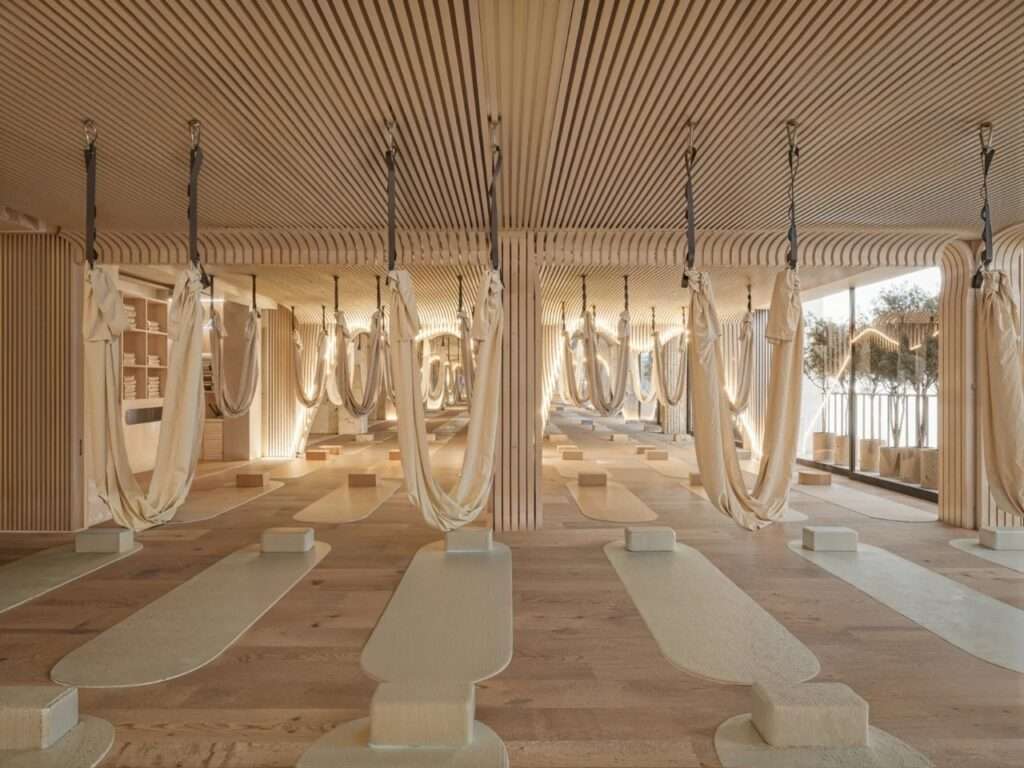 Aerial yoga hammocks suspended from a slatted oak timber ceiling over yoga mats in a spacious studio room.