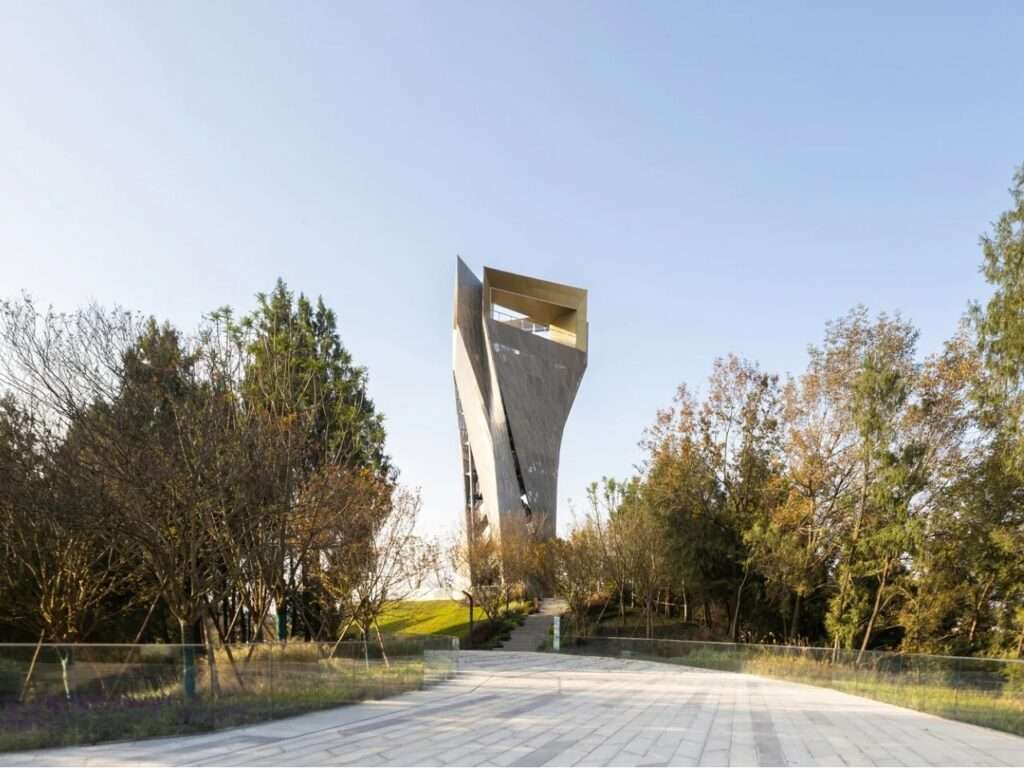 Vertical observation tower rising above a wooded park along a paved approach path.