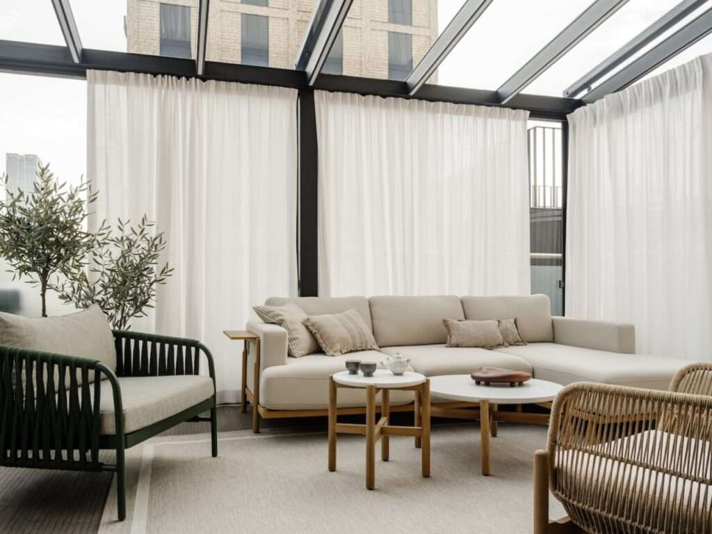 Glass-roof sunroom lounge with beige sectionals, woven armchairs, round marble coffee tables, and floor-to-ceiling sheer white curtains.