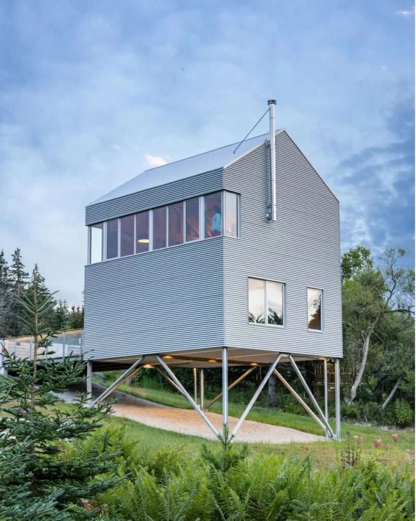 Angle view of the elevated Skybox house on steel pilotis showcasing corrugated metal siding, upper window band, and steel chimney flue.