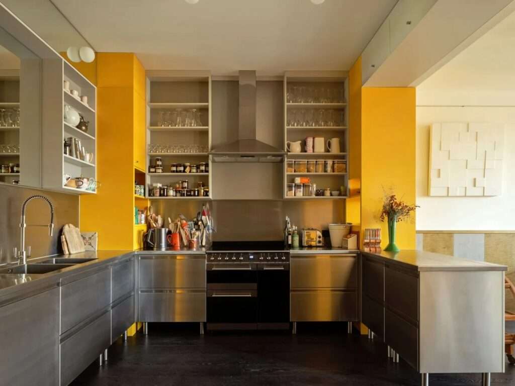 Industrial stainless steel kitchen counters paired with vibrant yellow accent cabinets and open shelving.