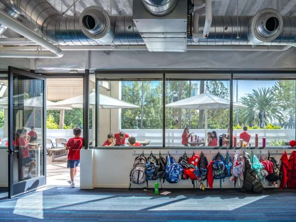 Student backpacks hanging on wall hooks below a interior window looking out onto an outdoor patio dining area with patio umbrellas.