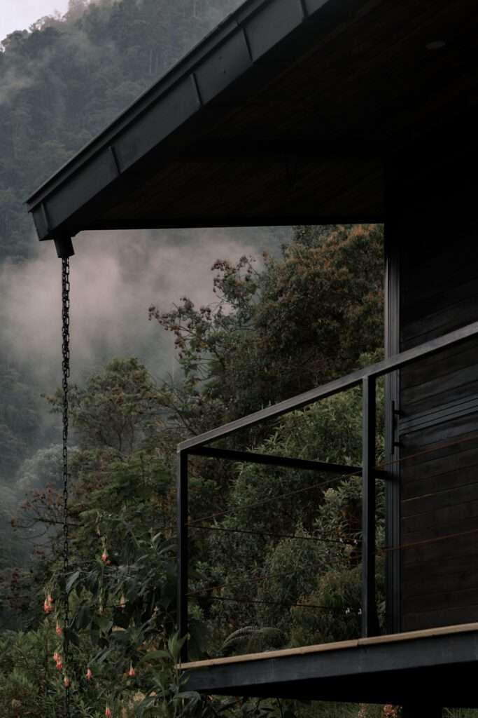 Close-up of dark wooden cladding, a cantilevered steel-railed balcony, and a rain chain hanging beneath a misty canopy.