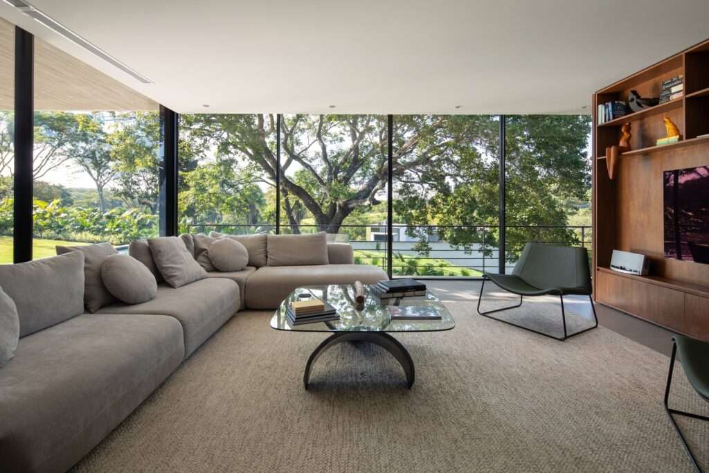 Modern open-plan living room in Casa Galería featuring a grey sectional sofa, floor-to-ceiling glass walls, and scenic views of surrounding treetop canopy.