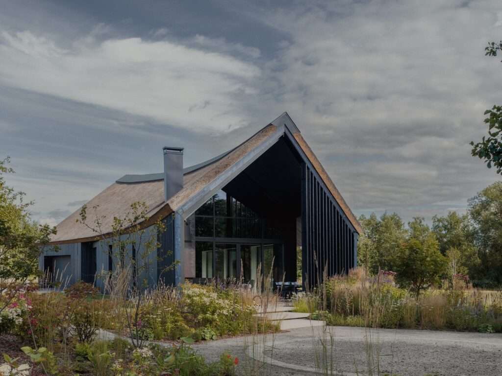 Large gabled residential structure with dark vertical timber cladding, thatched roof, and double-height glass facade surrounded by wildflowers.