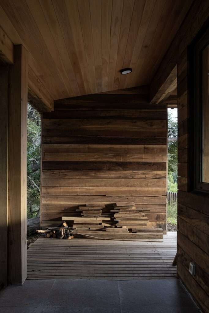 Sheltered entrance portal clad in dark horizontal wood planks with stone flooring and stacked firewood at Casa El Trébol.