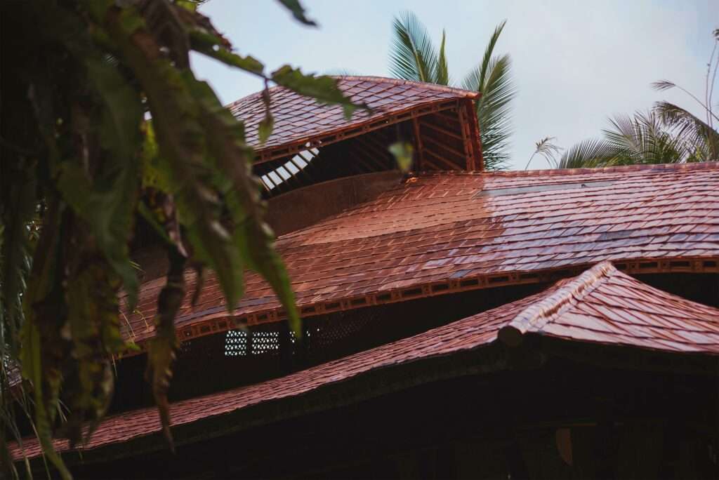 Close-up exterior view of the multi-tiered terracotta tile roof of the Hideout Leaf cabin, showing the overlap of tiles and the bamboo structural edge beam against a background of palm leaves.
