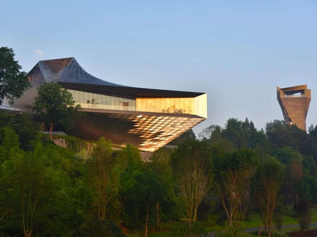 Side view of Chengdu Pavilion perched on a green hill with the vertical observation tower in the background at sunset.