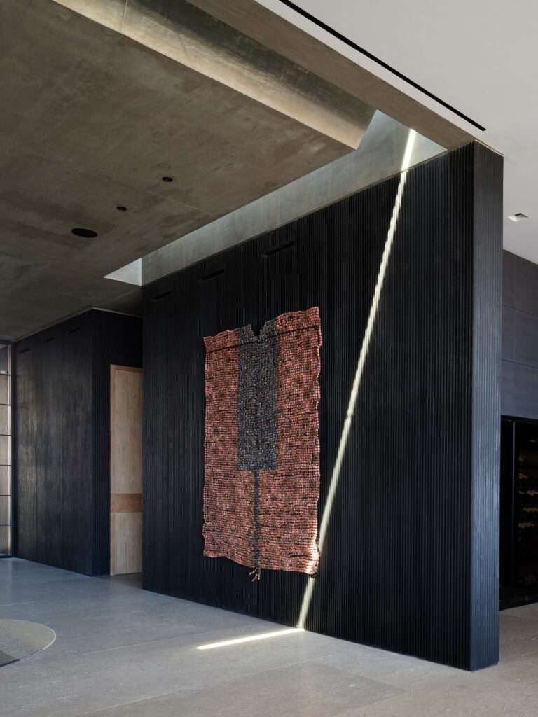 Interior hallway featuring a narrow skylight casting a beam of light over dark fluted concrete wall and woven textile artwork.
