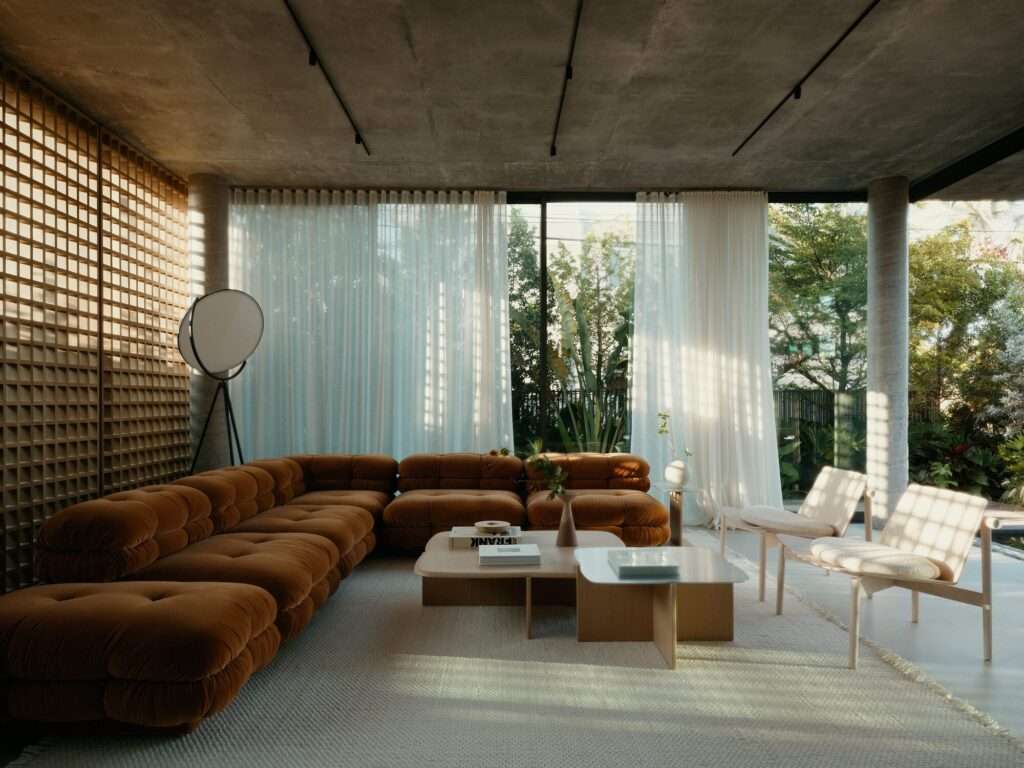 Interior shot of a sunlit living room with a brown velvet sectional sofa, sheer white curtains, and a square wooden lattice wall screen.