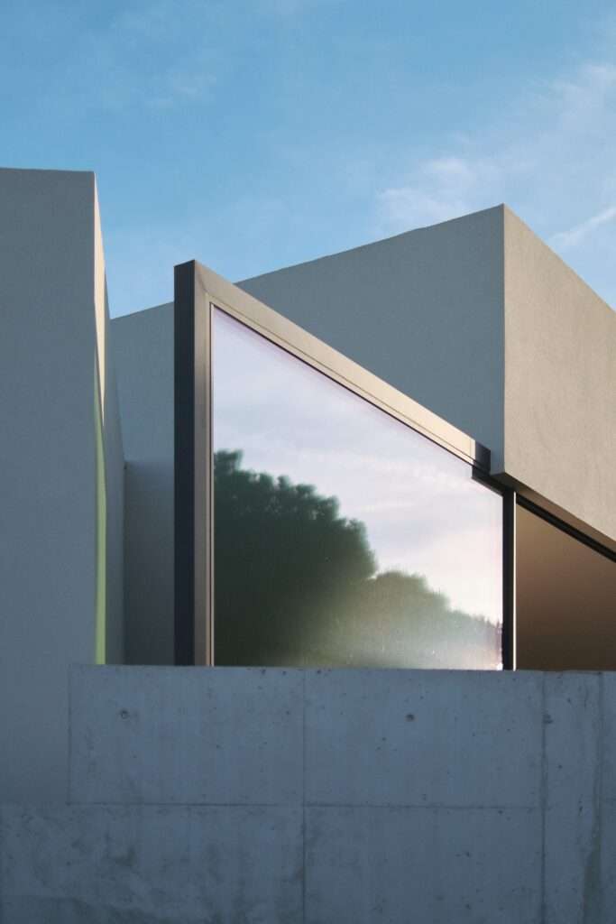 Close-up architectural shot of a triangular glass window and dark metal frame set into white facade walls above a textured concrete retaining wall, reflecting the sky and tree silhouettes at sunset.