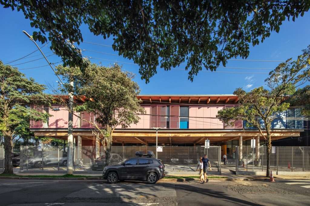 Exterior facade view of the school building with red louvers, timber posts, street trees, security fencing, and a perimeter street crosswalk.