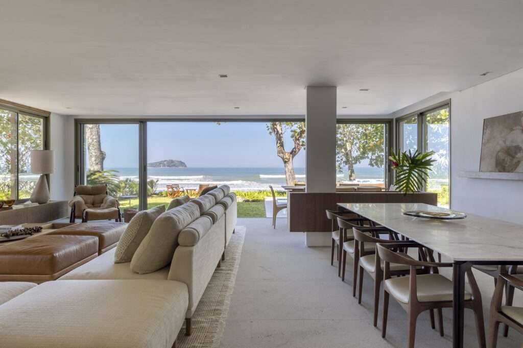 Bright open-plan interior of Casa ADS with a dining table, beige sectional sofa, and floor-to-ceiling glass doors opening to a beach and ocean panorama.
