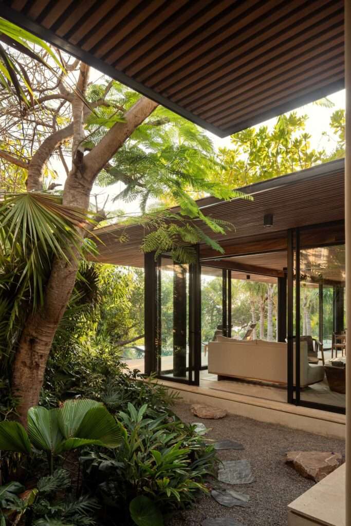 View from a lush garden looking through large open glass doors into a modern living room and out toward a sunlit terrace.