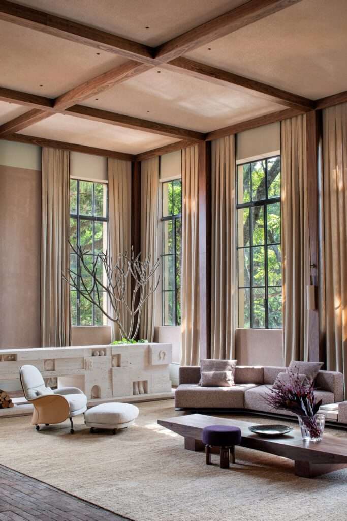Vertical view of a sunlit living room featuring tall curtained windows, exposed wooden ceiling beams, and a sculpted stone fireplace.