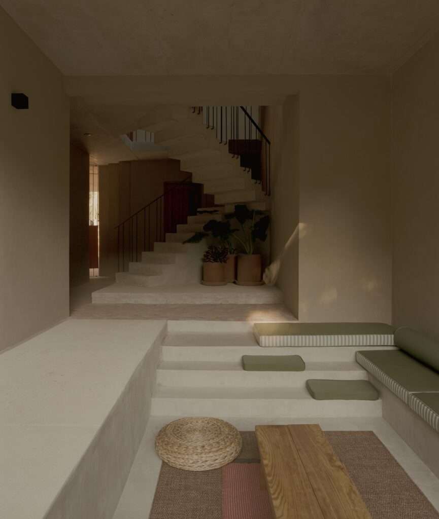 Interior entry lounge with concrete stepped seating, woven floor mat, wooden table, and a spiral staircase visible in the background.