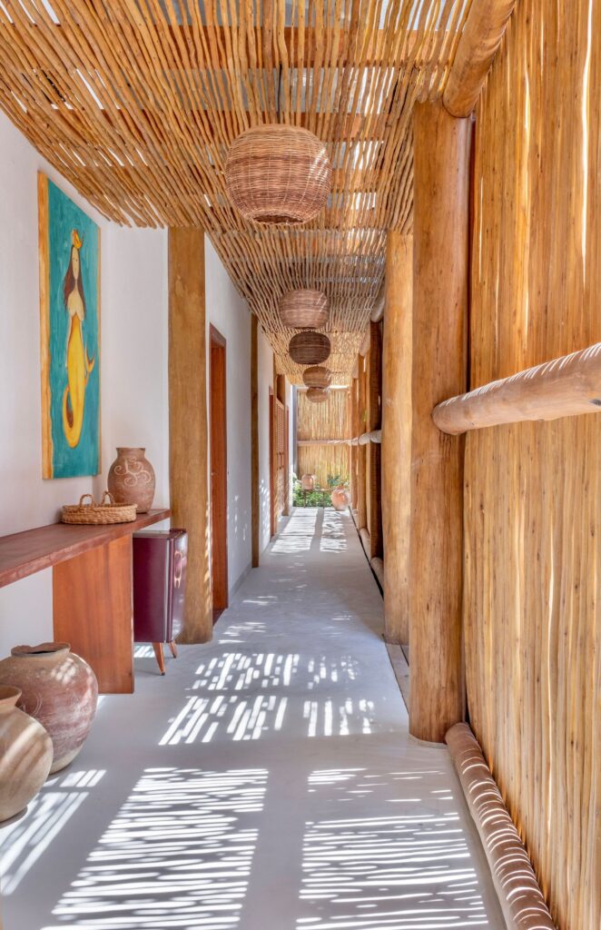Perspective view of a long residential hallway with biriba branch ceiling, hanging woven pendant lights, and dappled sunlight cast on the floor.