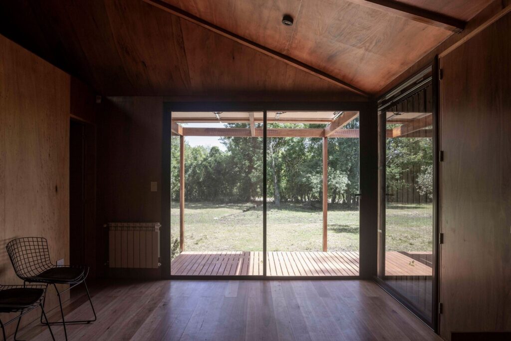 Warm wood-paneled living space inside Etchegoyen House featuring large sliding glass doors opening onto an outdoor wooden deck surrounded by trees.
