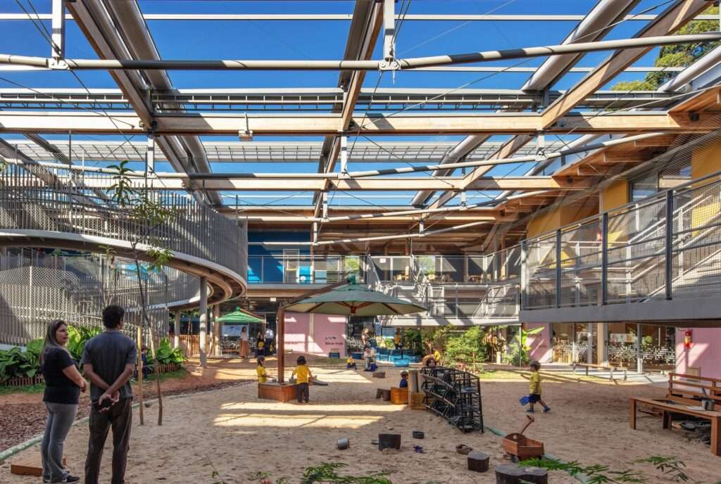 Ground-level perspective of the central sandy courtyard with children playing under parasols, framed by timber beams and steel mesh balustrades.