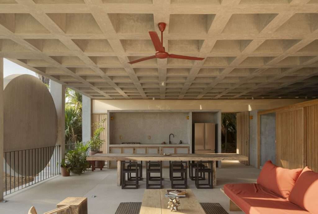 Open dining and kitchen area with a red ceiling fan, long wooden table, concrete kitchen niche, and coffered slab overhead.
