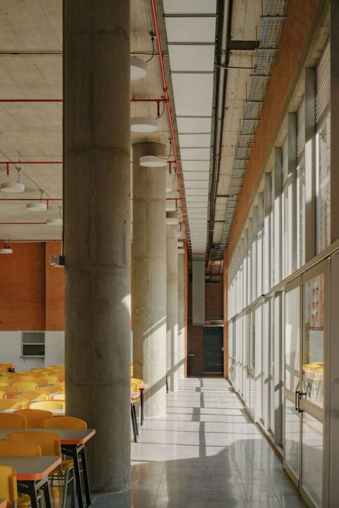 Sunlit interior dining hall with circular concrete columns, floor-to-ceiling glass walls, and exposed ceiling conduits.