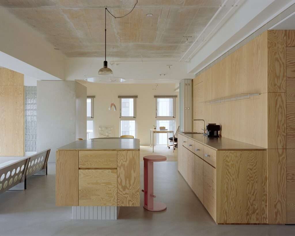 Perspective view through kitchen and corridor of Raw and Warm Residence showing pine plywood counter, island, exposed concrete ceiling, pink stool, and background windows.