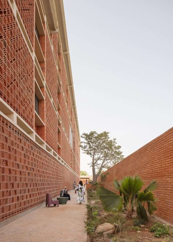 Linear ground-floor walkway flanked by a tall terracotta mashrabiya facade on one side and a red brick garden wall on the other.