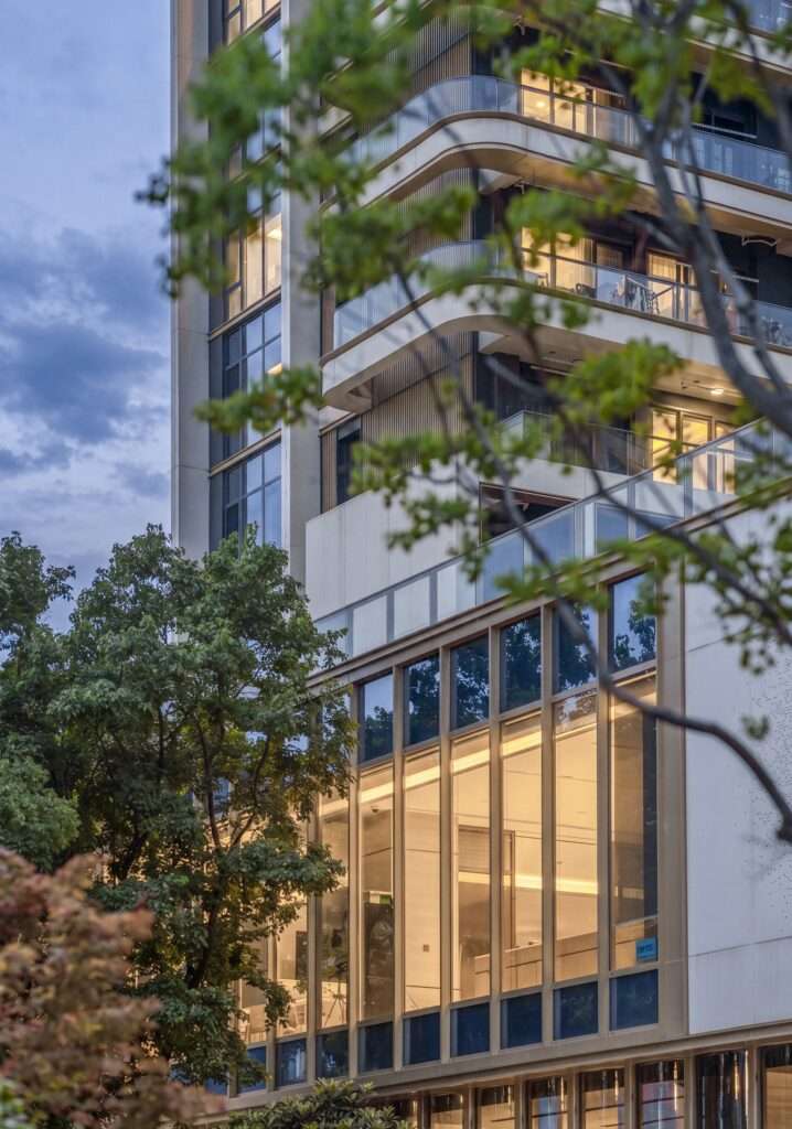 Modern building facade at dusk featuring curved upper balconies and a brightly illuminated double-height glass ground floor curtain wall.