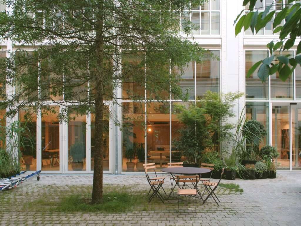 Outdoor dining table and chairs in a green courtyard garden set against large glass windows of the renovated office building.