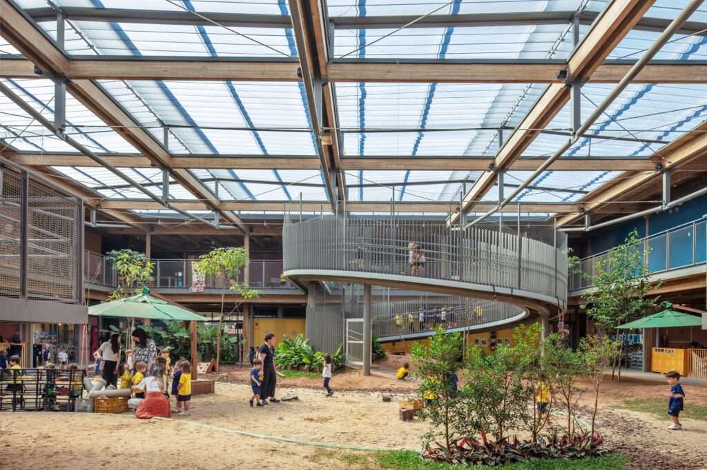Wide shot of children and teachers interacting in the sandy courtyard beneath a translucent roof, wooden frame, and curved access ramp.