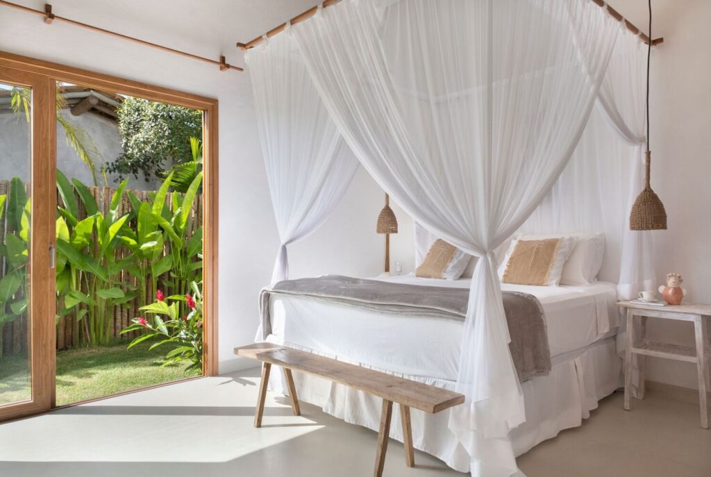 Bright bedroom suite featuring a white canopy bed with mosquito netting and glass sliding doors opening to a private tropical garden.