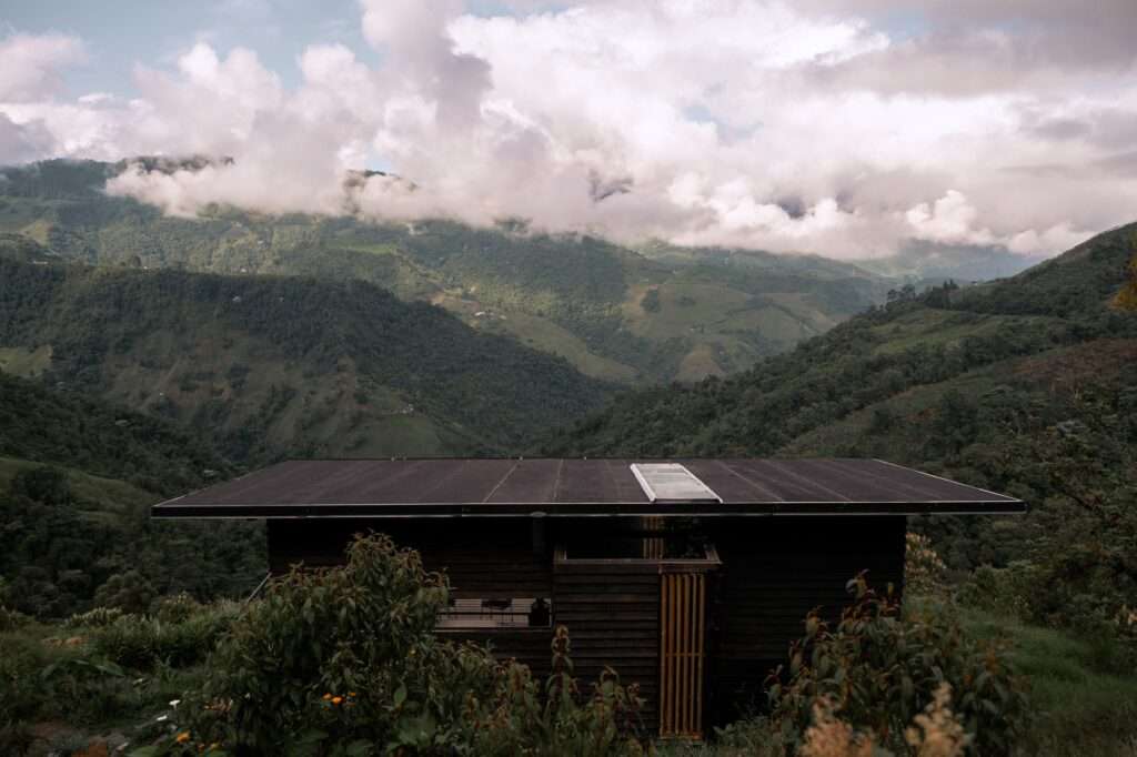 Elevated view looking down at a flat dark roof with a skylight, surrounded by vast green mountain ranges under cloudy skies.