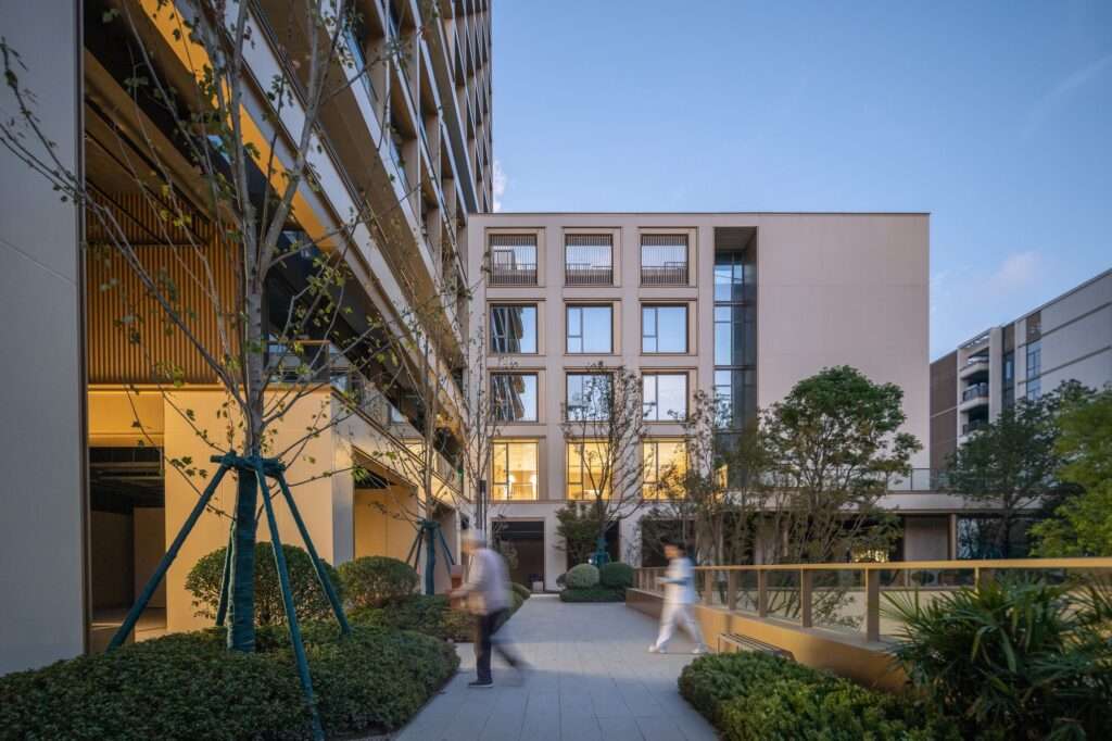 Elevated outdoor walkway and terrace garden between residential blocks with residents and staff walking at dusk amidst planted trees and warm lighting.