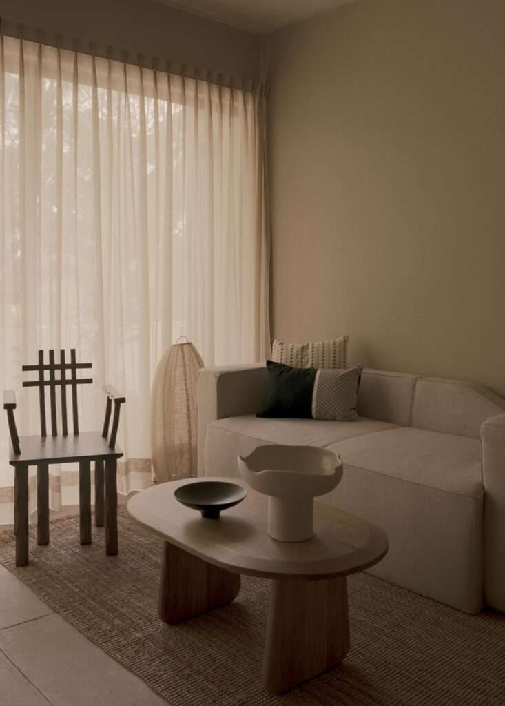 Minimalist living room with beige fabric sofa, wooden coffee table, dark accent chair, and sheer curtains draped over wide windows.