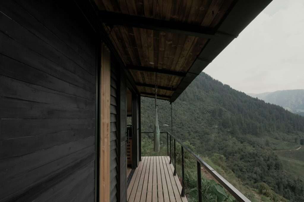 Side perspective of a dark wooden cabin corridor and wooden deck balcony featuring a hammock facing dense green slopes.