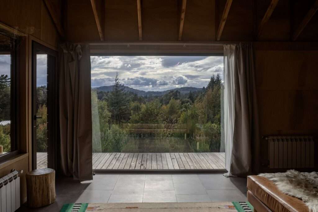 Large square glass window framing green mountain forest scenery from inside a warm timber-clad room.