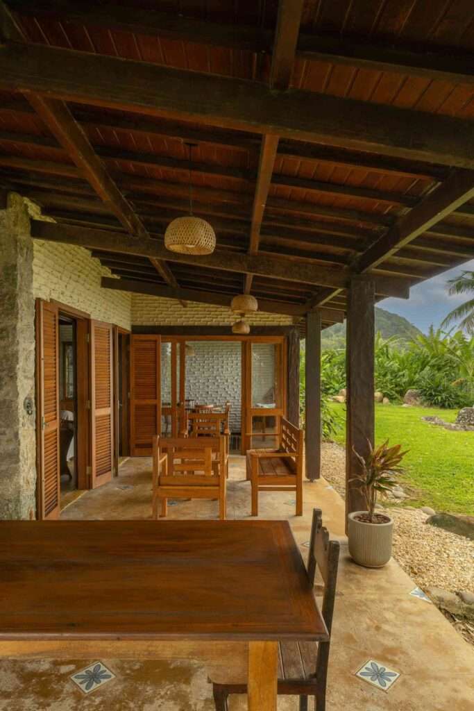 Covered veranda patio with wooden furniture, woven pendant light fixtures, exposed timber ceiling beams, and stone wall detailing.
