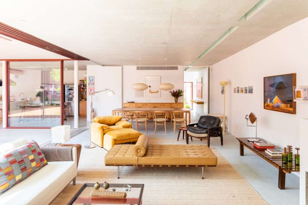 Spacious contemporary interior showing a dining table with ten chairs, yellow armchairs, a leather daybed, and sliding glass walls facing the garden.