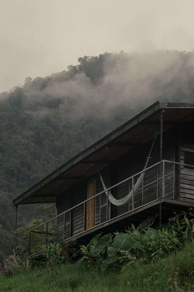 Low-angle view of a dark timber shelter elevated above a grassy hill with a hammock on the balcony under mist-covered trees.
