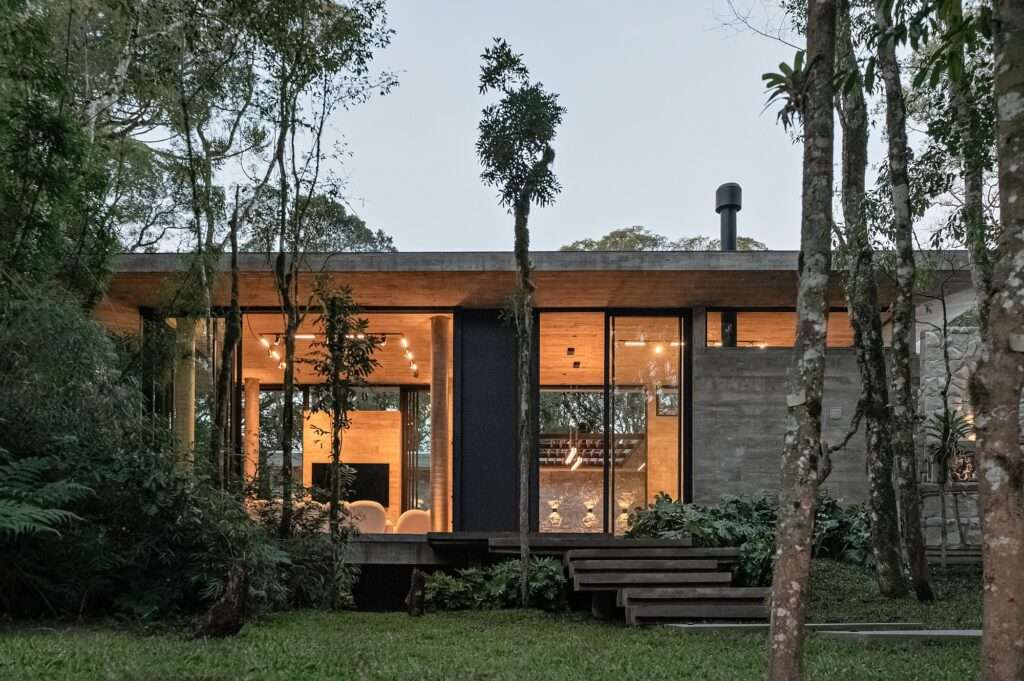 Dusk exterior view of Casa de Vidro illuminated from within, showcasing an open social space and central fireplace structure.