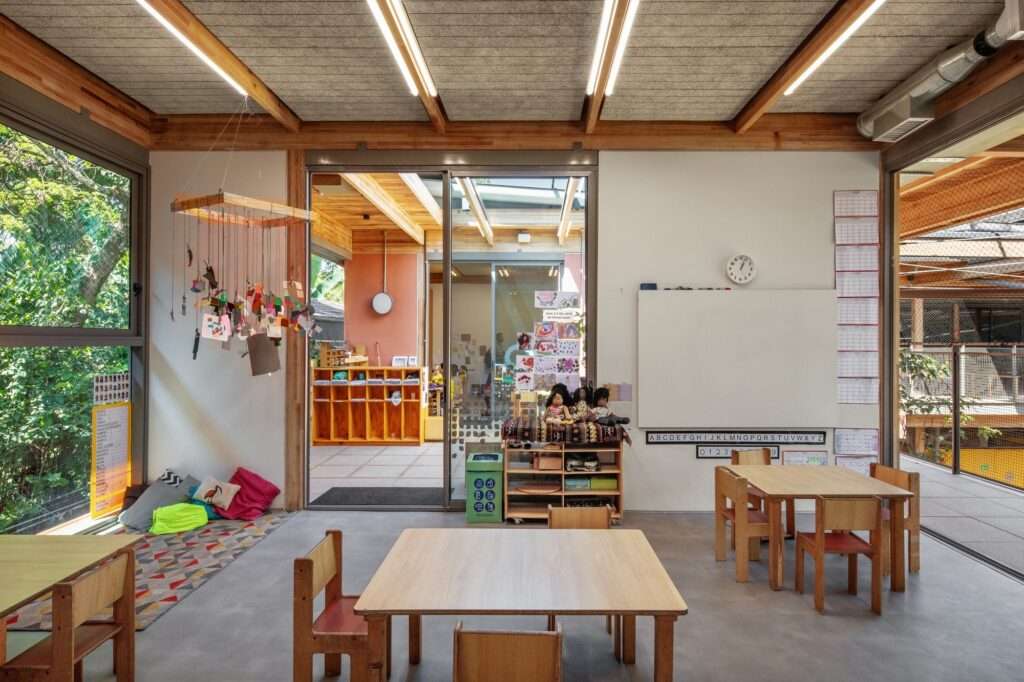 Kindergarten classroom interior with timber ceiling beams, acoustic ceiling panels, wooden children furniture, whiteboard, and sliding glass door to courtyard.