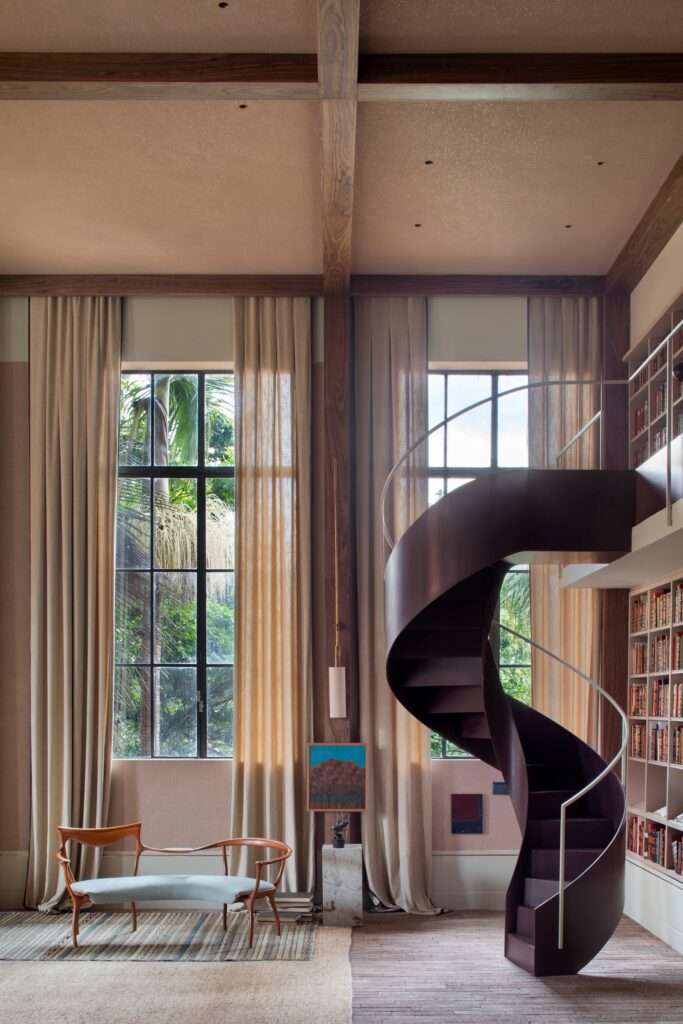 Dark purple spiral staircase alongside tall glass windows with sheer curtains and a classic wooden lounge chair.