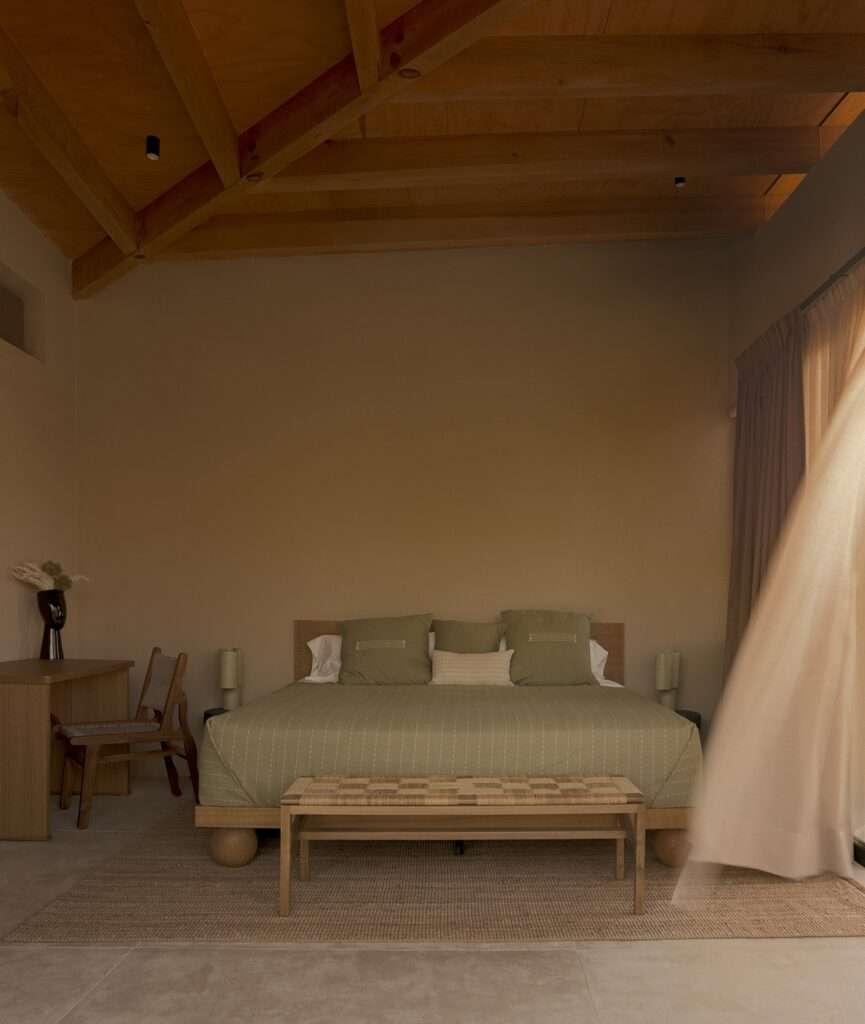 Bedroom interior with exposed wooden ceiling beams, king bed with green linens, woven bench, and sheer white curtains blowing in the wind.