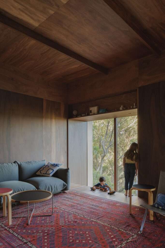 An internal view of EA House’s living area with two children playing with toy cars near a large window, framed by the dark plywood interior.