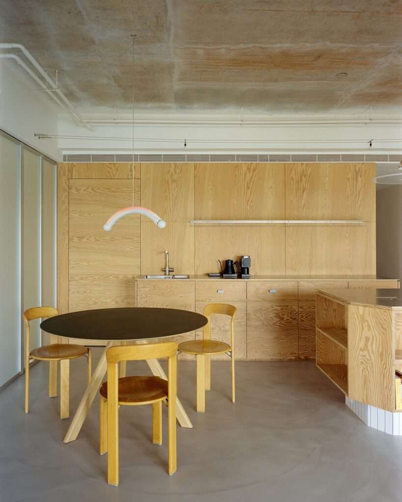 Dining area in Raw and Warm Residence featuring round table with dark top, wooden chairs, playful arc pendant lamp, pine plywood wall cabinets, and exposed concrete ceiling.