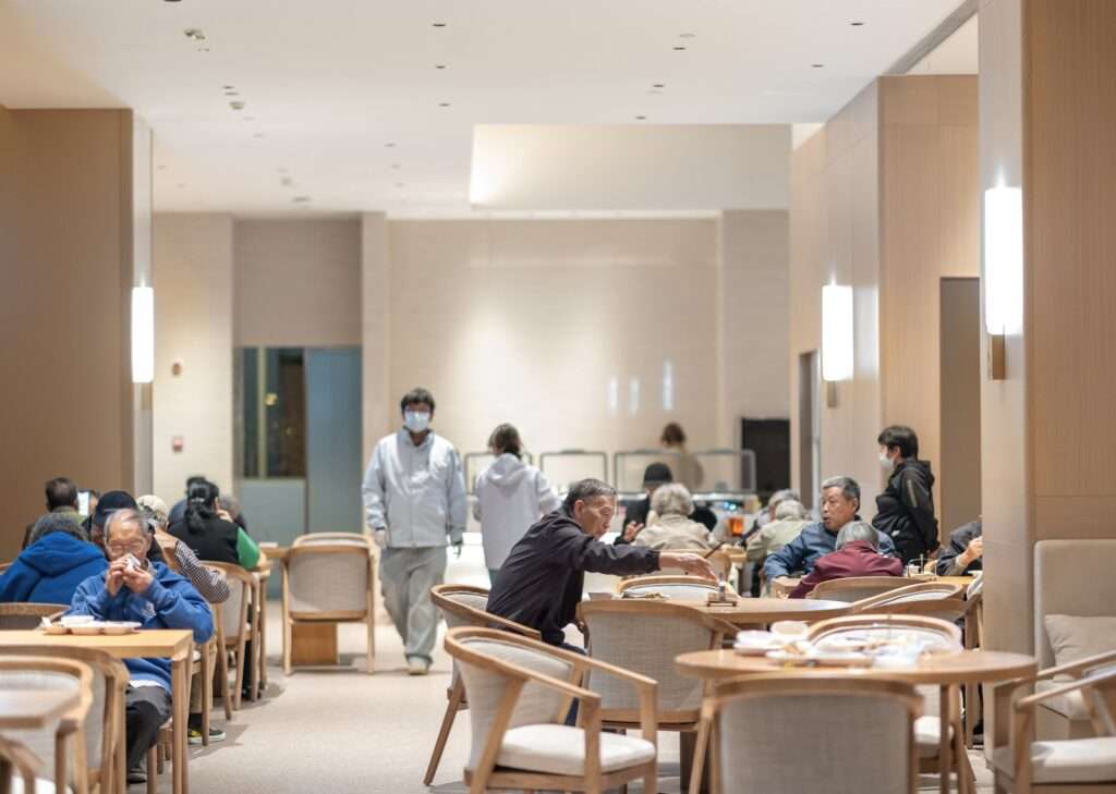 Senior residents dining together at warm wooden tables in a spacious, well-lit cafeteria dining hall.