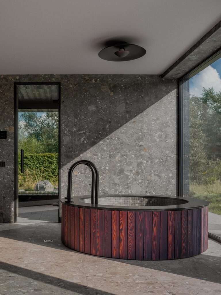 Circular wooden hot tub placed on terrazzo tile floor with large window overlooking garden and dark grey stone walls.